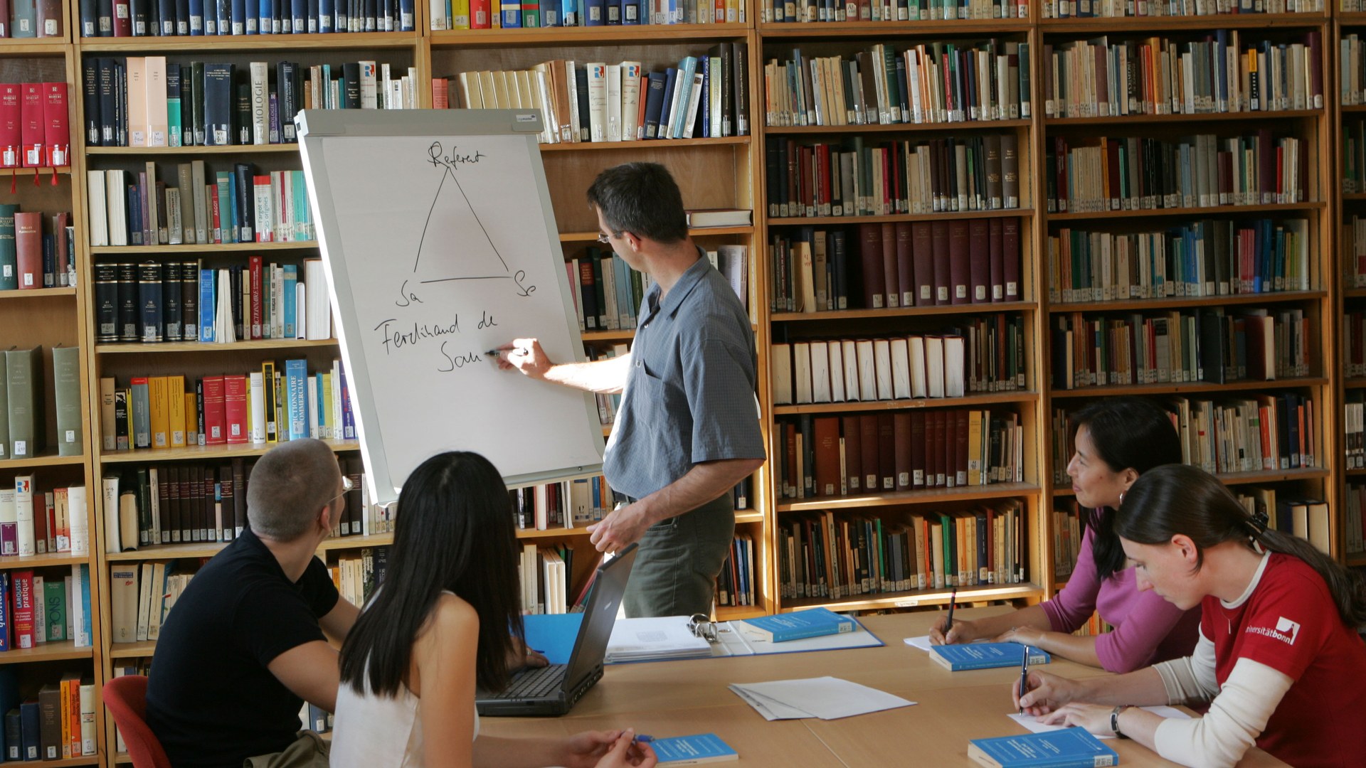 Eine Wissenschaftlerin und ein Wissenschaftler arbeiten hinter einer Glasfassade und mischen Chemikalien mit Großgeräten.